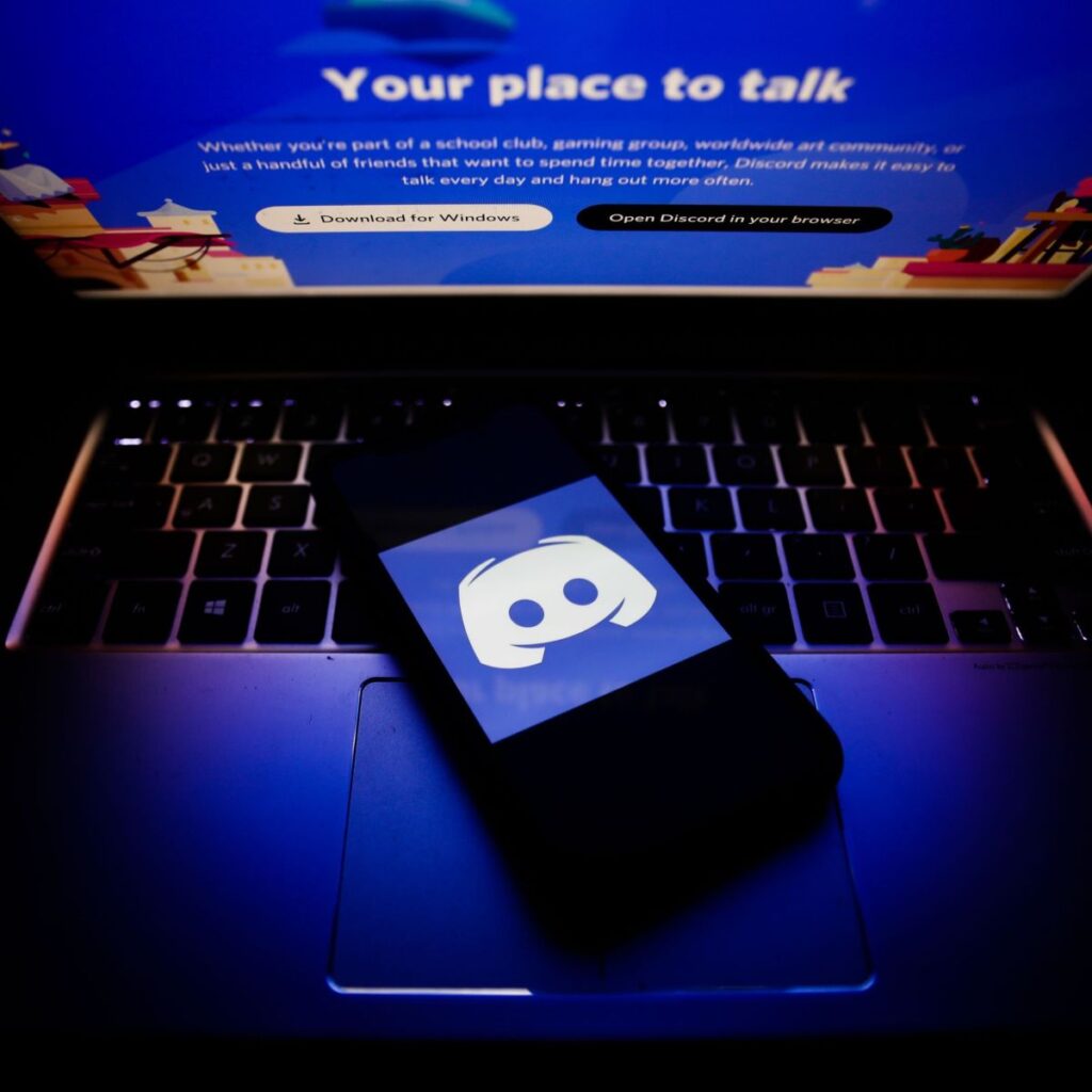 Smartphone com o logotipo do Discord sobre um notebook exibindo a página inicial da plataforma.