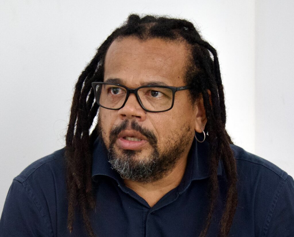 Kleber Rosa, homem negro de dreadlocks e óculos, veste camisa azul escura e fala durante entrevista.