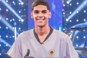 O influenciador Luva de Pedreiro sorrindo, vestindo camisa cinza com emblema, em estúdio com luzes ao fundo.