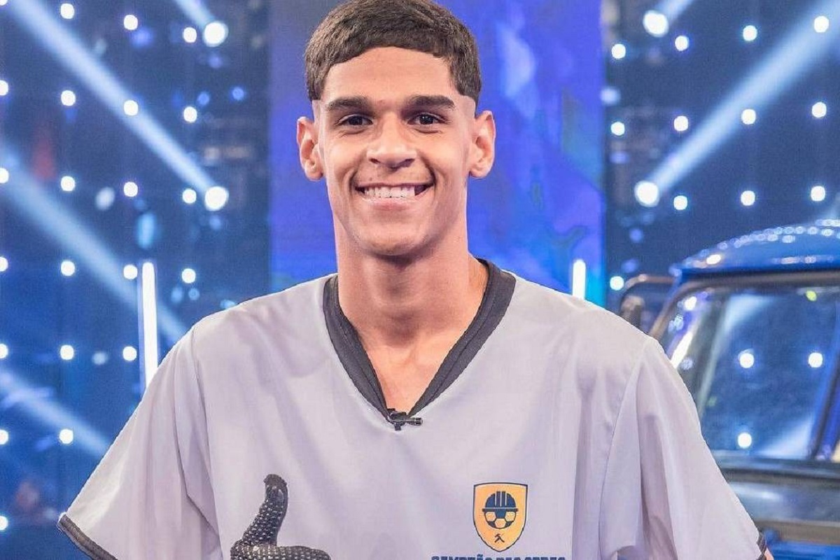 O influenciador Luva de Pedreiro sorrindo, vestindo camisa cinza com emblema, em estúdio com luzes ao fundo.