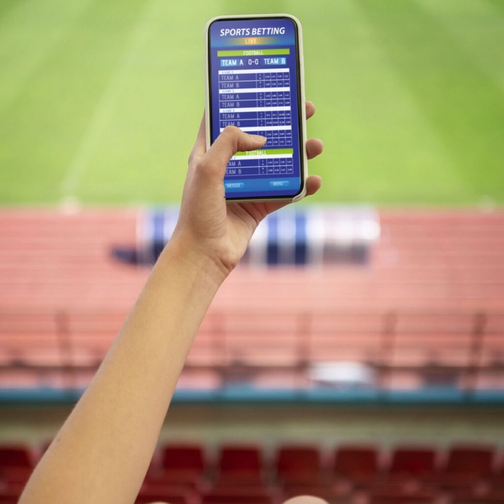 Mão segurando smartphone com aplicativo de apostas esportivas em um estádio de futebol.