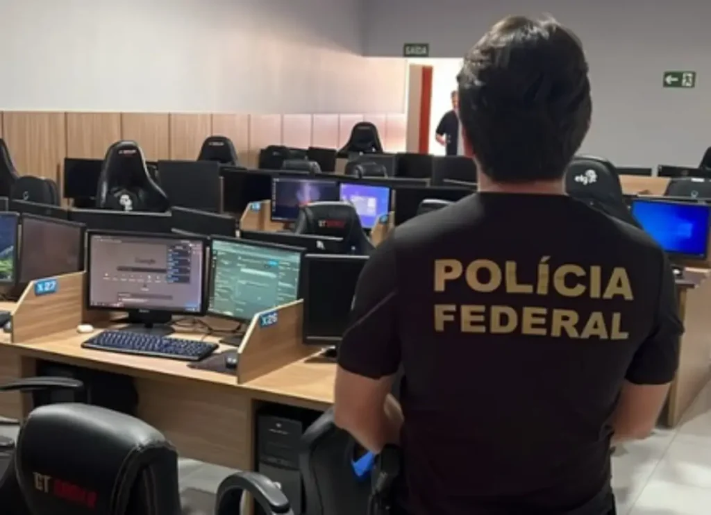 Agente da Polícia Federal observa escritório com diversas estações de trabalho equipadas com computadores.