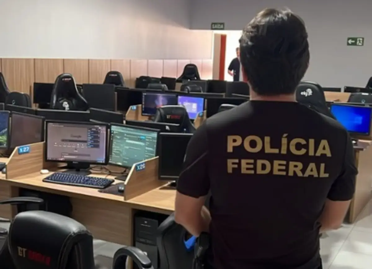 Agente da Polícia Federal observa escritório com diversas estações de trabalho equipadas com computadores.