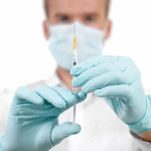 Profissional de saúde de máscara e luvas segurando uma seringa fina em primeiro plano.